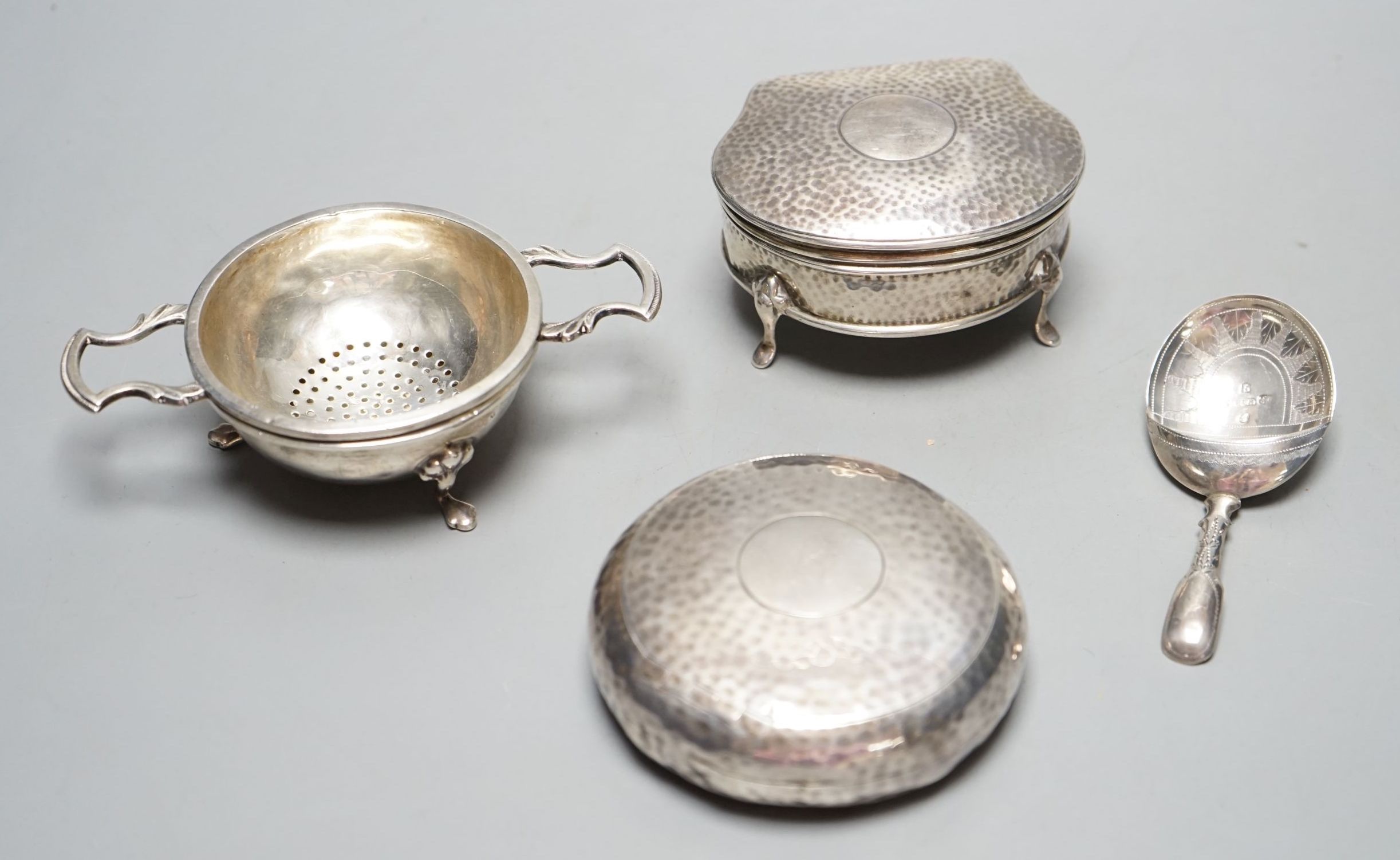 An Edwardian planished silver circular tobacco box, by George Unite, Birmingham, 1902, 79mm, a similar silver trinket box, a George III silver caddy spoons, Birmingham, 1814 and a modern silver teas strainer on stand.